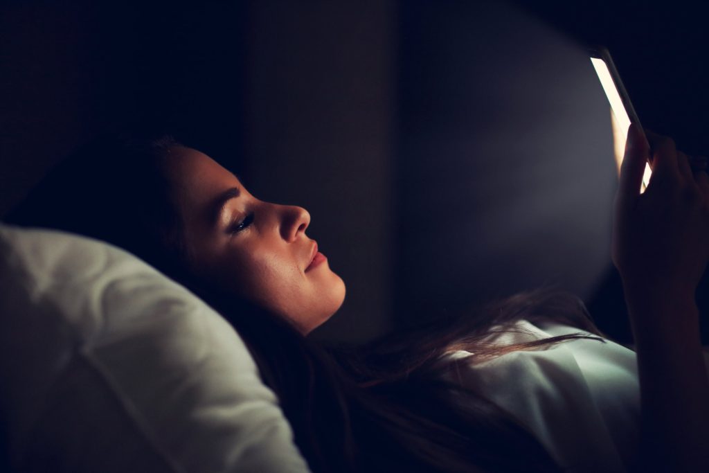 A young woman scrolling on her smartphone while trying to fall asleep in bed