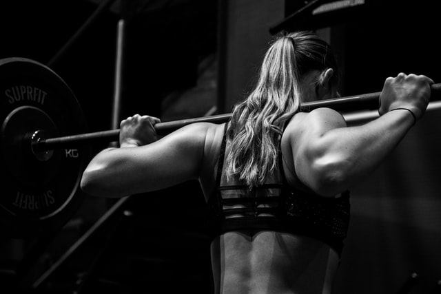 A black-and-white image of a strong woman holding a barbell above her shoulders getting ready to squat in the gym